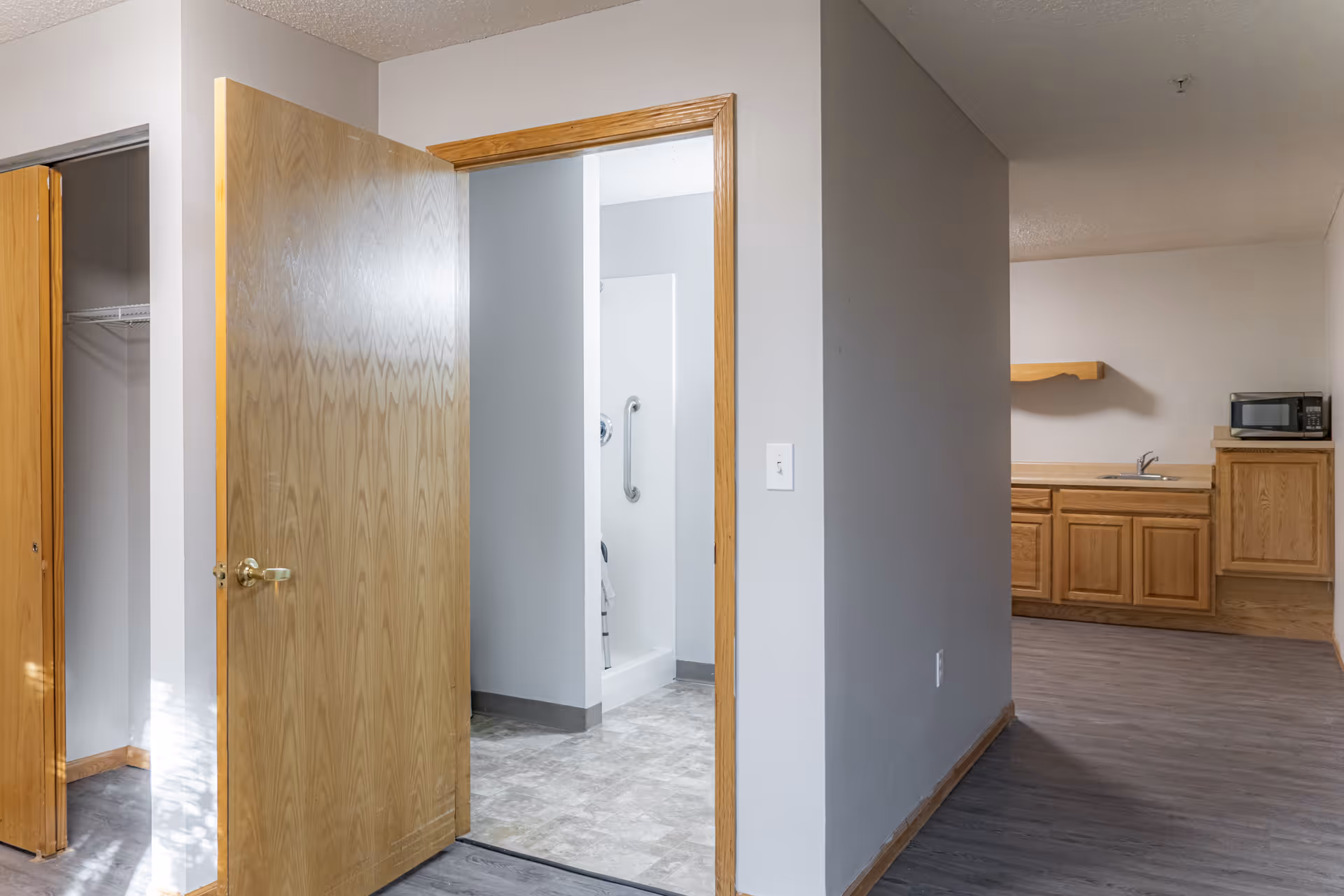 Interior view of a small apartment with an open wooden door revealing an accessible bathroom and a kitchenette with wooden cabinets and a microwave.