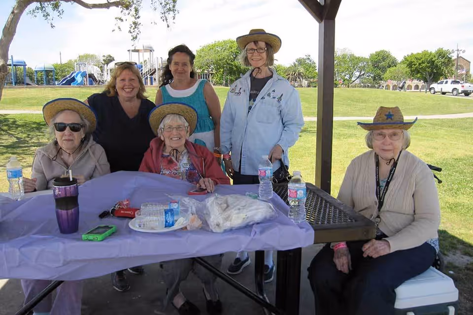 Five elderly women and one middle-aged woman gathered under a shaded pavilion at a park. Three of the elderly women are seated at a picnic table covered with a purple tablecloth, water bottles, and other items. Two elderly women and one middle-aged woman stand behind the table. Some of the elderly women are wearing straw hats. In the background, there is a playground with slides and green grassy areas.