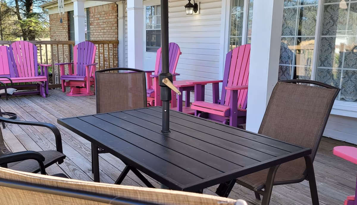Outdoor wooden deck with a black metal table and several brown mesh chairs. In the background, there are bright pink Adirondack chairs lined up against a white building with windows and a brick wall section. The deck is surrounded by a wooden railing and trees are visible in the distance.