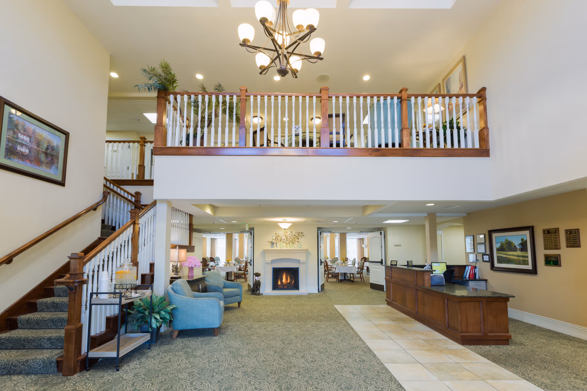 Spacious assisted living facility lobby with a green patterned carpet and tiled walkway. There is a wooden reception desk on the right, two blue armchairs and a small table with a lamp and flowers on the left. A staircase with wooden handrails and white balusters leads to an upper balcony area with similar railing. A chandelier hangs from the ceiling, and a fireplace with a fire burning is centered at the back wall. Dining areas are visible through doorways behind the fireplace.