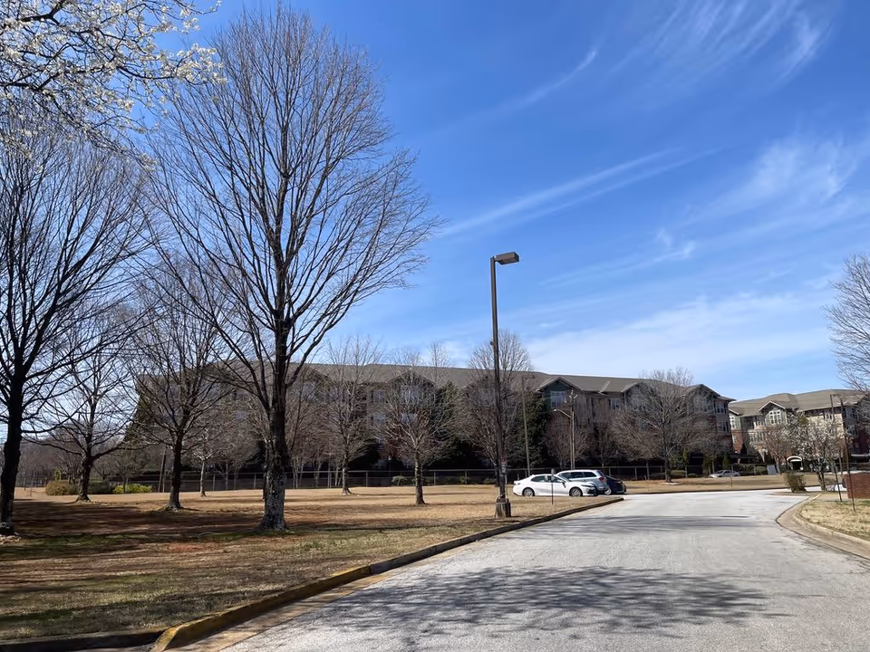 Driveway and parking area leading to a multi-story senior living building with leafless trees under a blue sky.