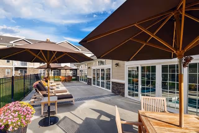 Outdoor patio area at Summit at Mill Hill with large umbrellas, wooden tables and chairs, cushioned lounge chairs, potted flowers, and a view of the building exterior under a partly cloudy sky.