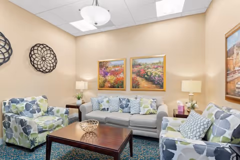 A cozy living room with a gray sofa and two floral-patterned armchairs arranged around a dark wooden coffee table. The walls are beige and decorated with two framed paintings of colorful flower gardens and a decorative metal wall piece. There are two table lamps on side tables beside the sofa, and the floor is covered with a blue patterned carpet.