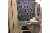 Bathroom with a bathtub and blue-gray patterned shower curtain, toilet, pedestal sink with mirror, and two dark bath mats on a tiled floor.
