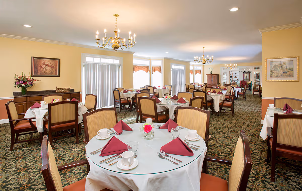 A spacious dining room in a senior living facility with multiple round tables covered with white tablecloths and glass tops. Each table is set with white cups, saucers, silverware, and burgundy folded napkins. The room features patterned carpet, yellow walls, framed artwork, floral centerpieces, and brass chandeliers hanging from the ceiling. Large windows with vertical blinds and valances allow natural light to enter.