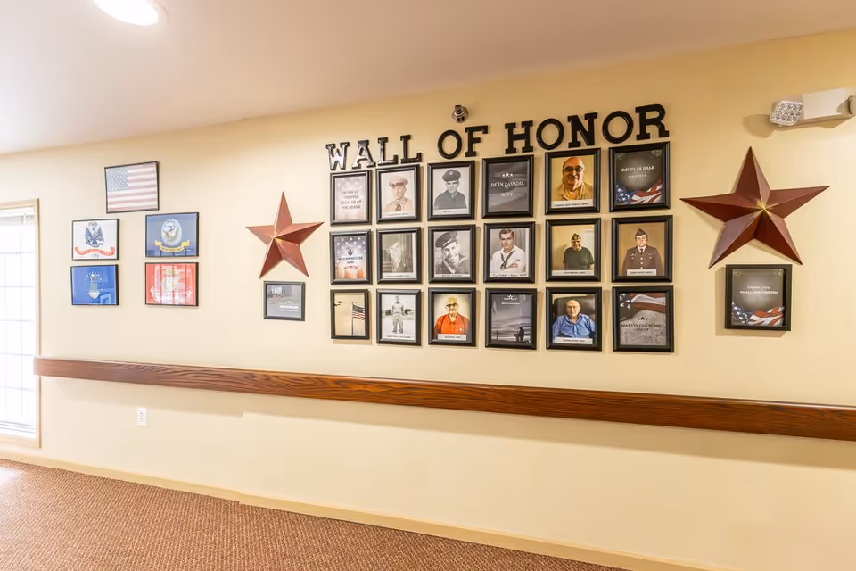 A hallway wall display titled 'Wall of Honor' featuring framed photographs and plaques honoring veterans, with two large decorative red stars and several military insignias and flags on the adjacent wall.