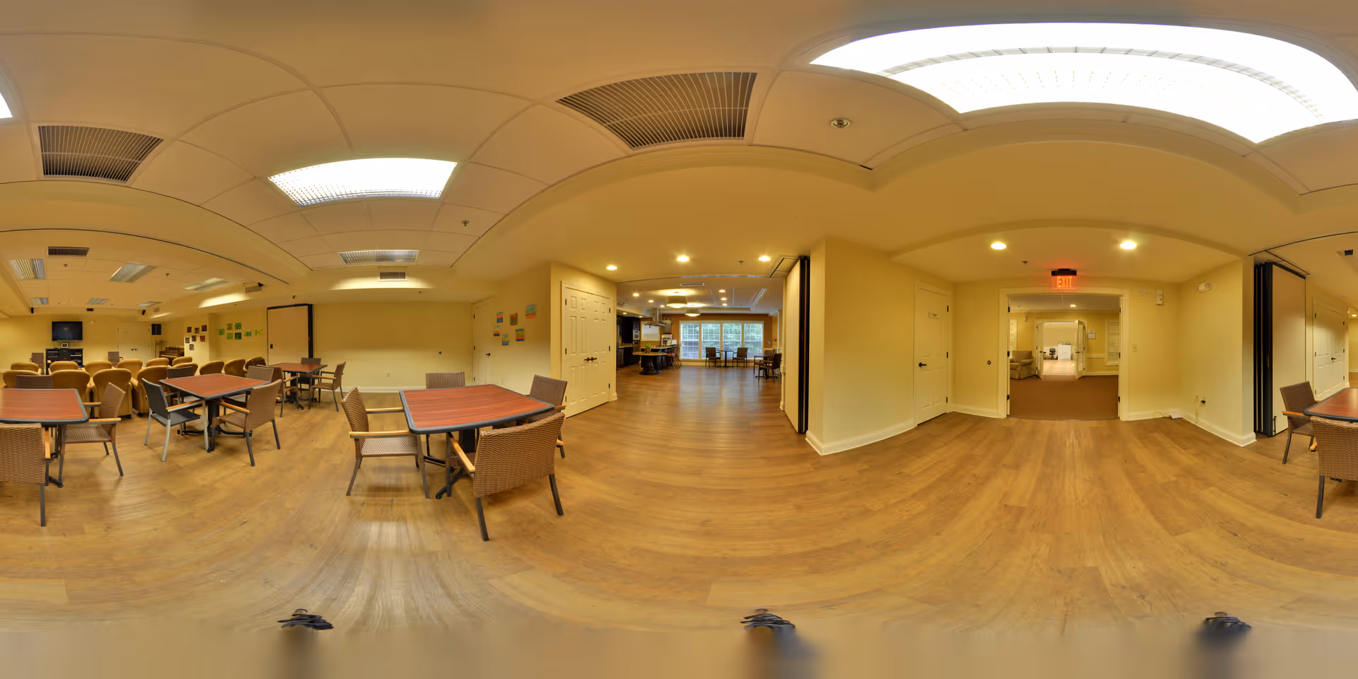 A spacious interior room with multiple tables and chairs arranged for group seating, featuring wood flooring and beige walls. The room is well-lit with ceiling lights and has a hallway leading to another area with more seating and large windows in the background.
