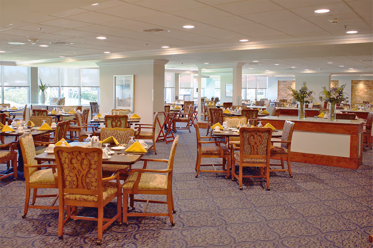 Spacious, well-lit dining room with multiple set tables, upholstered chairs, yellow napkins, and floral centerpieces.