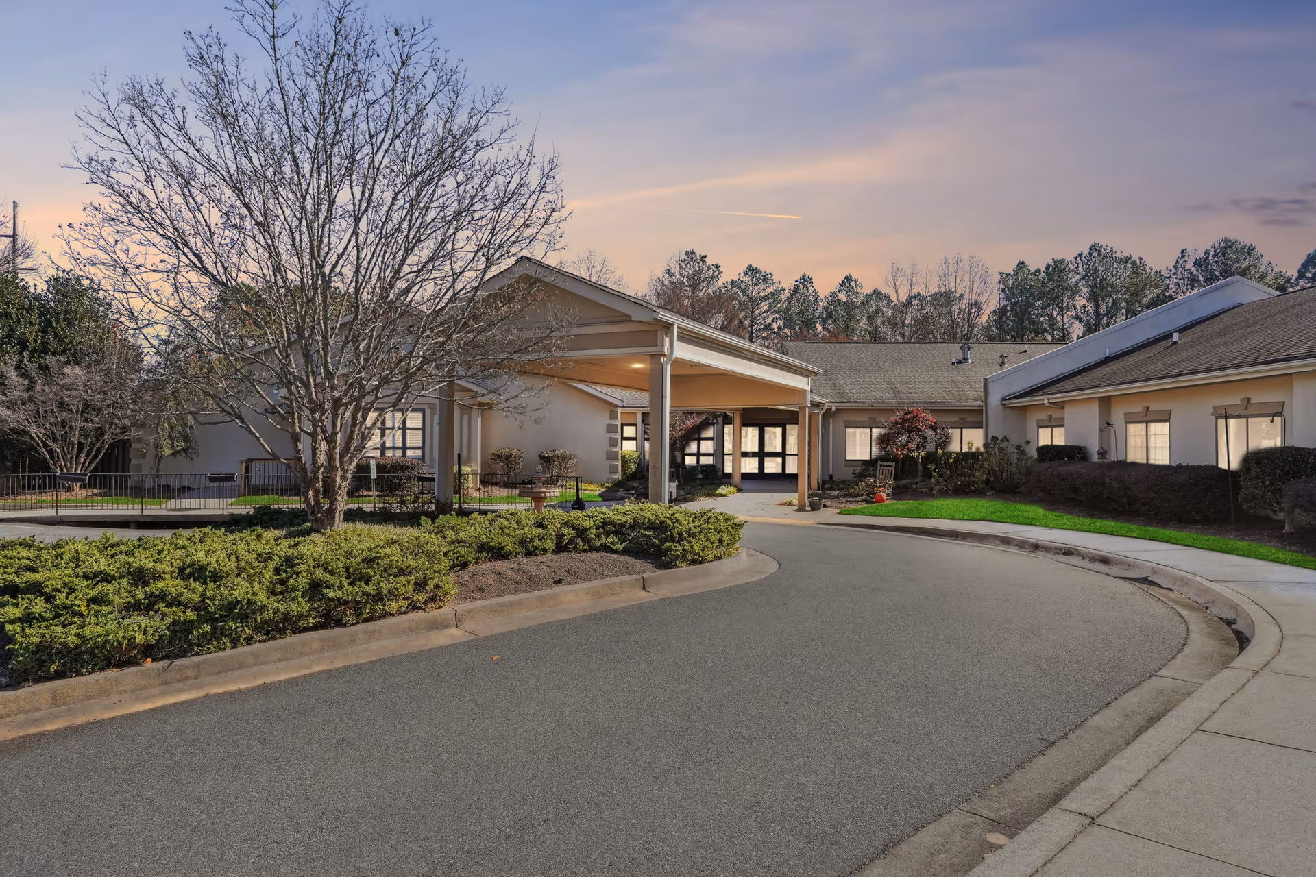 Exterior view of Cumming Health & Rehab facility at dusk, showing a curved driveway leading to a covered entrance with surrounding trees and shrubs.