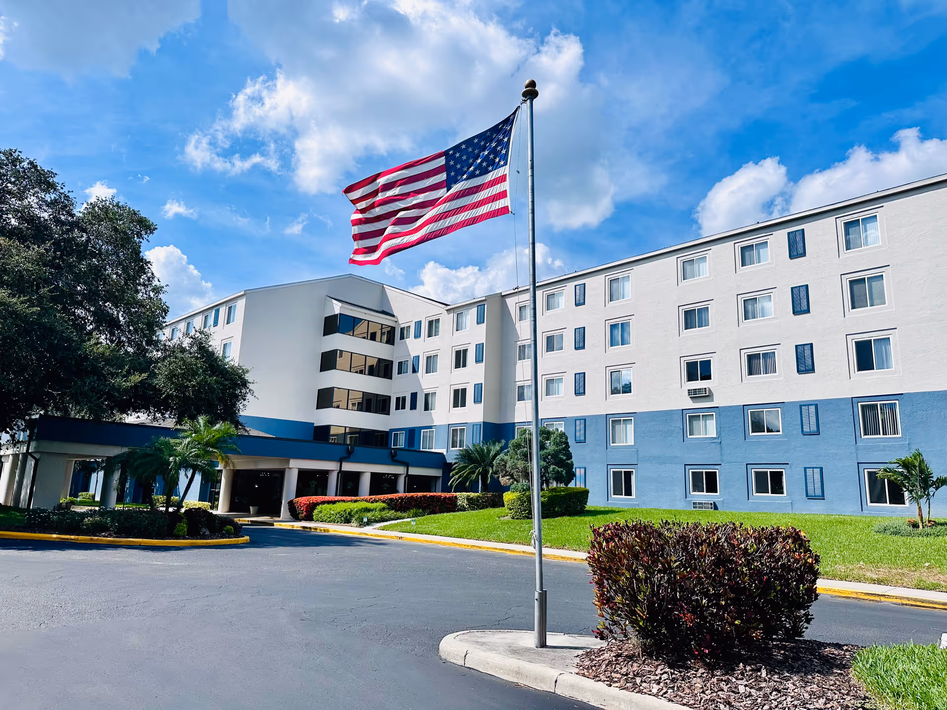 Front exterior of a multi-story white-and-blue senior living building with an American flag flying in front and landscaped grounds.
