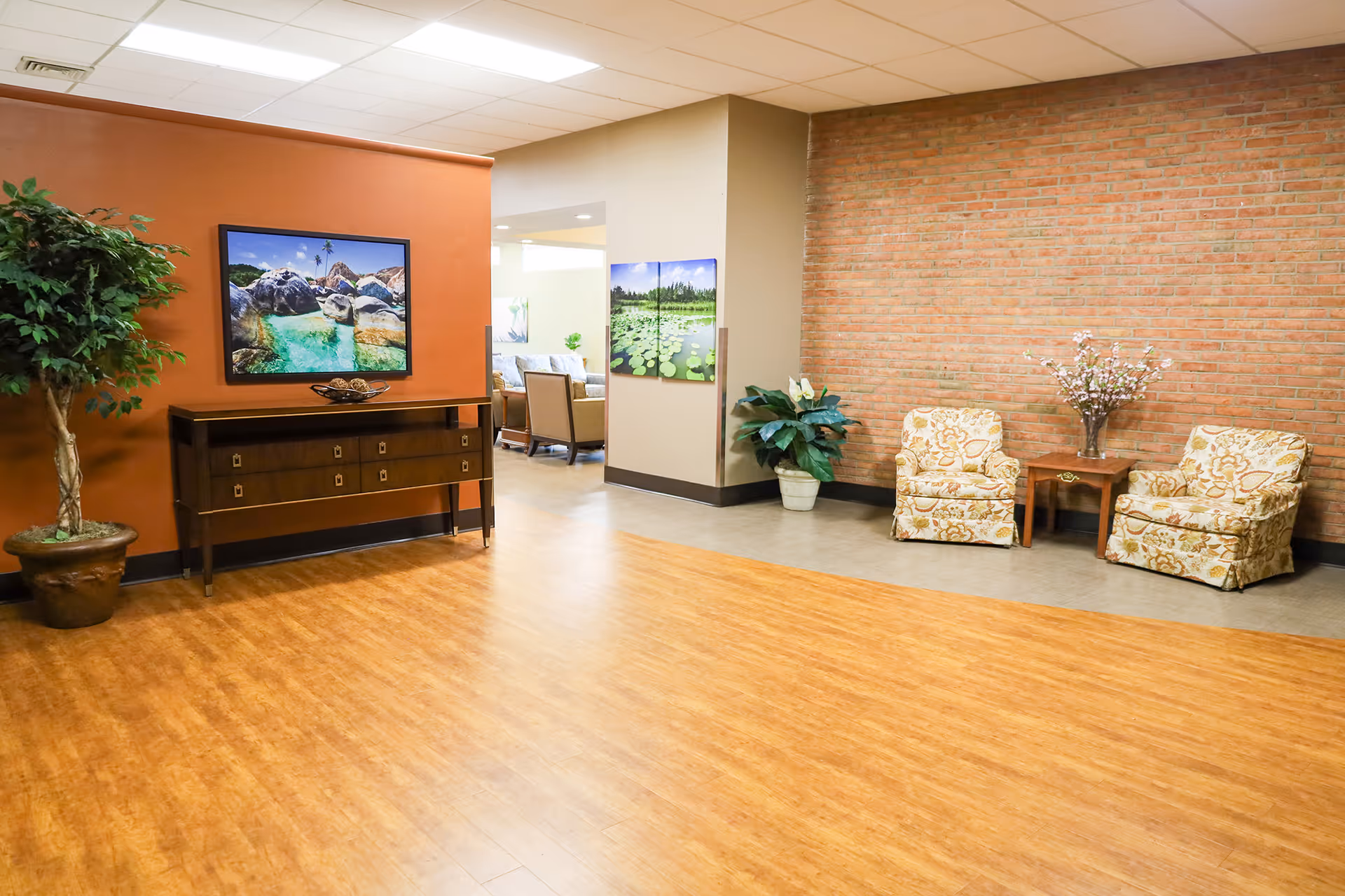 Open seating area in a senior living facility featuring wood floors, a console table with framed landscape art, potted plants, and two patterned armchairs beside a brick accent wall.