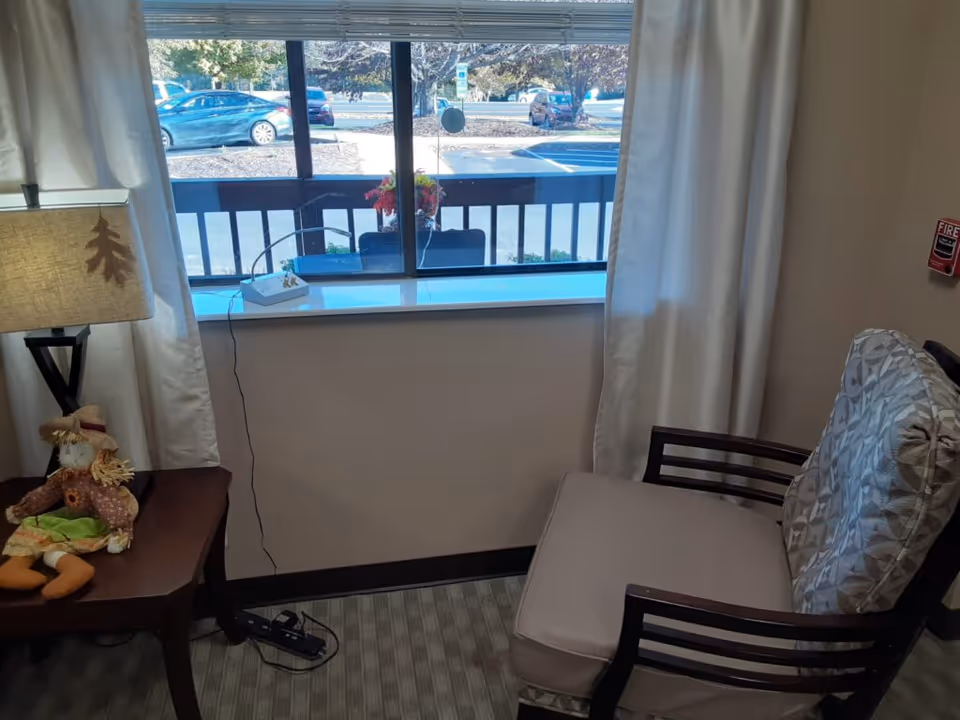 A cozy corner of a room with a cushioned chair and a side table. The table has a lamp with a leaf design on the shade and a small doll sitting next to it. A window with white curtains shows a view of parked cars and trees outside.