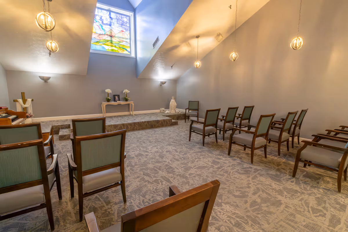 A small chapel or meditation room with several wooden chairs arranged in rows facing a raised platform. The platform has a small table with flowers and a framed picture, and a statue is placed to the side. A stained glass window depicting a landscape with mountains and clouds is above the platform. The room has soft lighting with hanging pendant lights and wall sconces.