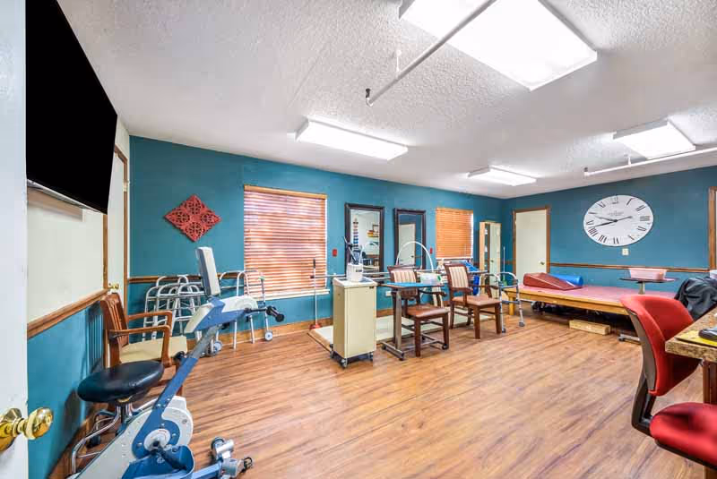 A bright therapy/exercise room with exercise equipment, walkers, chairs, and treatment tables against teal walls and wood flooring.