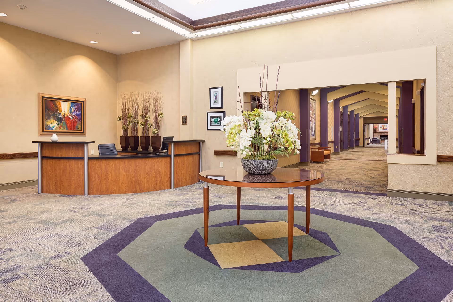 A spacious reception area with a wooden front desk on the left side, decorated with tall vases holding dried plants. In the center, there is a round wooden table with a large floral arrangement of white and green flowers. The floor features a patterned carpet with geometric shapes in shades of purple, green, and beige. The background shows a long hallway with purple pillars and additional seating areas.