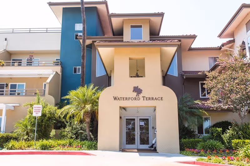 Front exterior view of Waterford Terrace Retirement Community building with a beige and teal facade, palm trees, and landscaped greenery around the entrance.