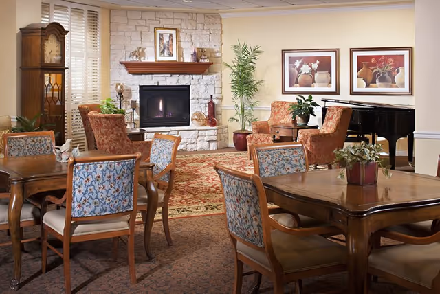 A cozy senior living common area with wooden tables and floral upholstered chairs, a stone fireplace with a lit fire, a grandfather clock, potted plants, framed floral artwork on the walls, and a black grand piano in the corner.