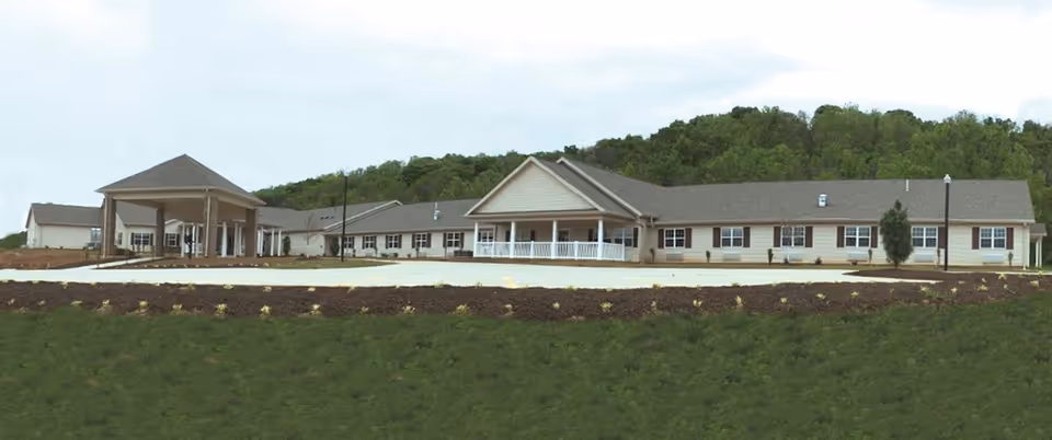 Exterior view of a single-story skilled nursing and rehabilitation facility with a covered entrance and a porch, surrounded by landscaped grounds and greenery in the background.
