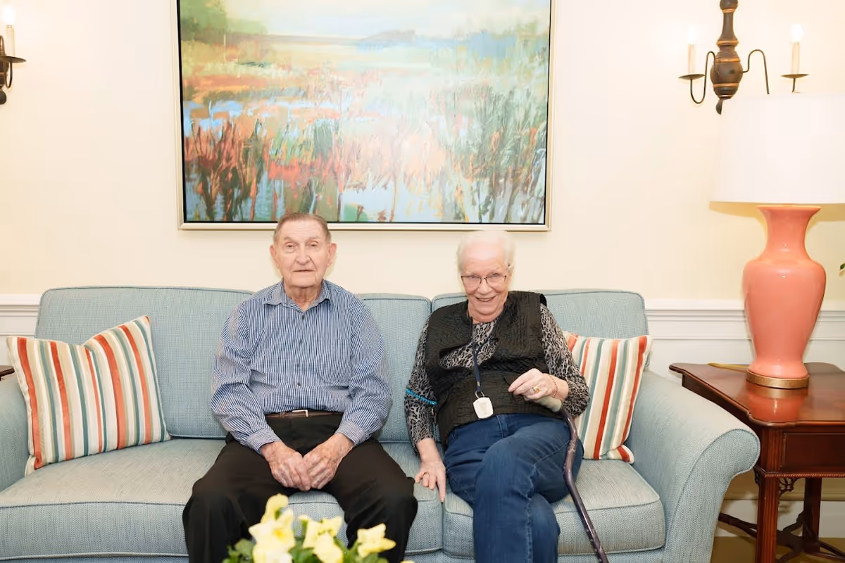 Two elderly individuals sitting on a light blue sofa in a well-lit room. The man on the left is wearing a striped shirt and dark pants, while the woman on the right is wearing glasses, a patterned top, and a black vest, holding a cane. Behind them is a colorful abstract painting, and on either side of the sofa are striped pillows. To the right is a wooden side table with a coral-colored lamp and a wall sconce with candles above it.