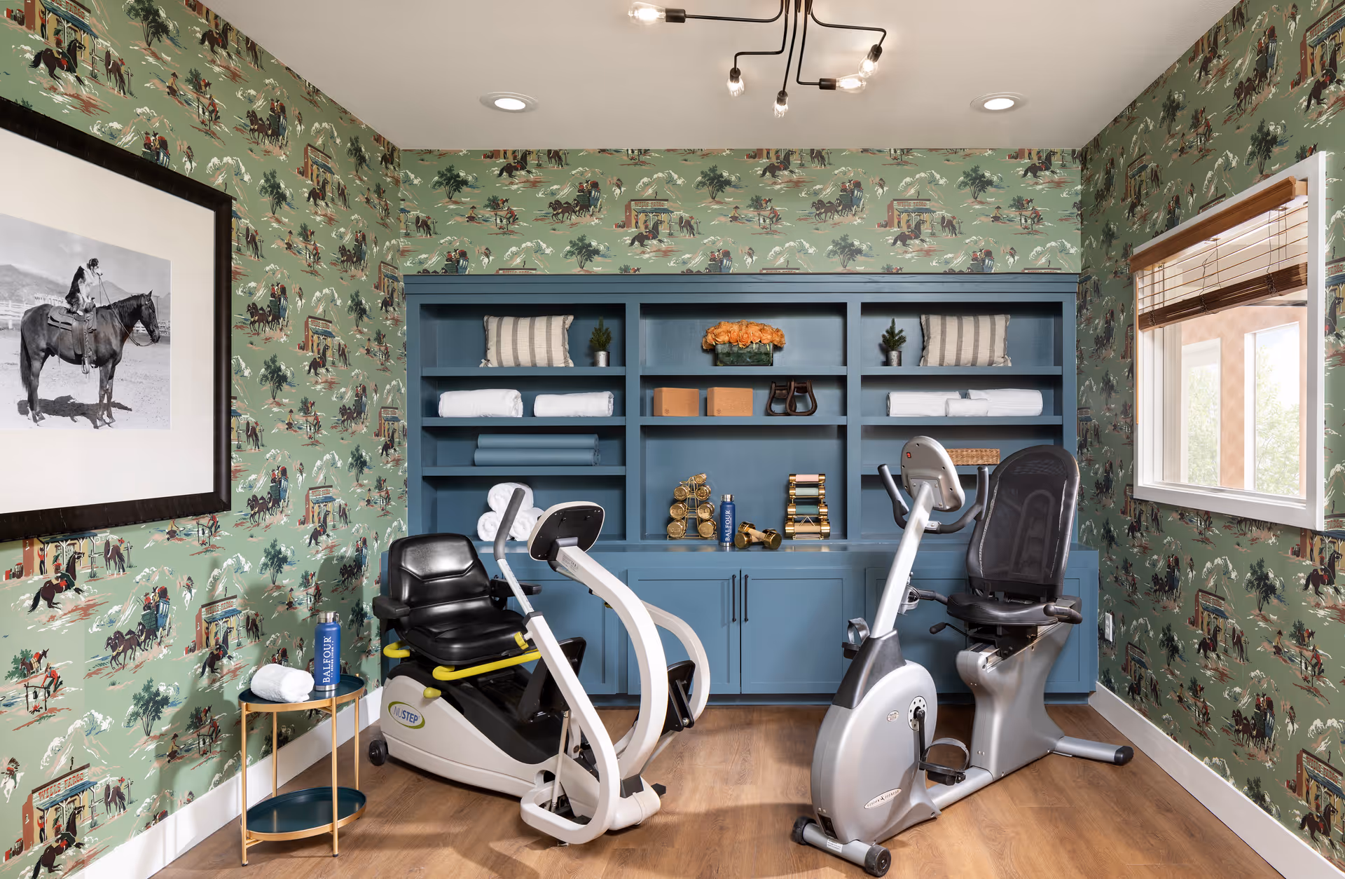 A small exercise room with two stationary recumbent bikes placed on a wooden floor. The walls are covered with green wallpaper featuring a vintage horse riding pattern. A blue built-in shelving unit holds folded towels, decorative pillows, plants, and small decorative items. A framed black and white photo of a person on a horse hangs on the left wall. A small round side table with a rolled towel and a water bottle is next to one of the bikes. A window with wooden blinds is on the right wall, allowing natural light into the room.