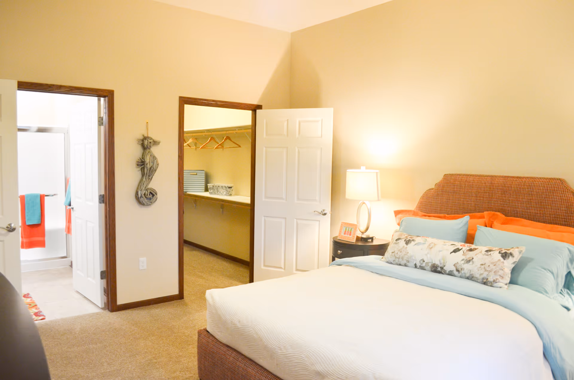 A bright bedroom with a neatly made bed featuring light blue and orange pillows and a floral decorative pillow. A bedside table with a lamp and a small framed picture is next to the bed. Two open doors reveal a walk-in closet with hangers and storage bins, and a bathroom with a shower and colorful towels.