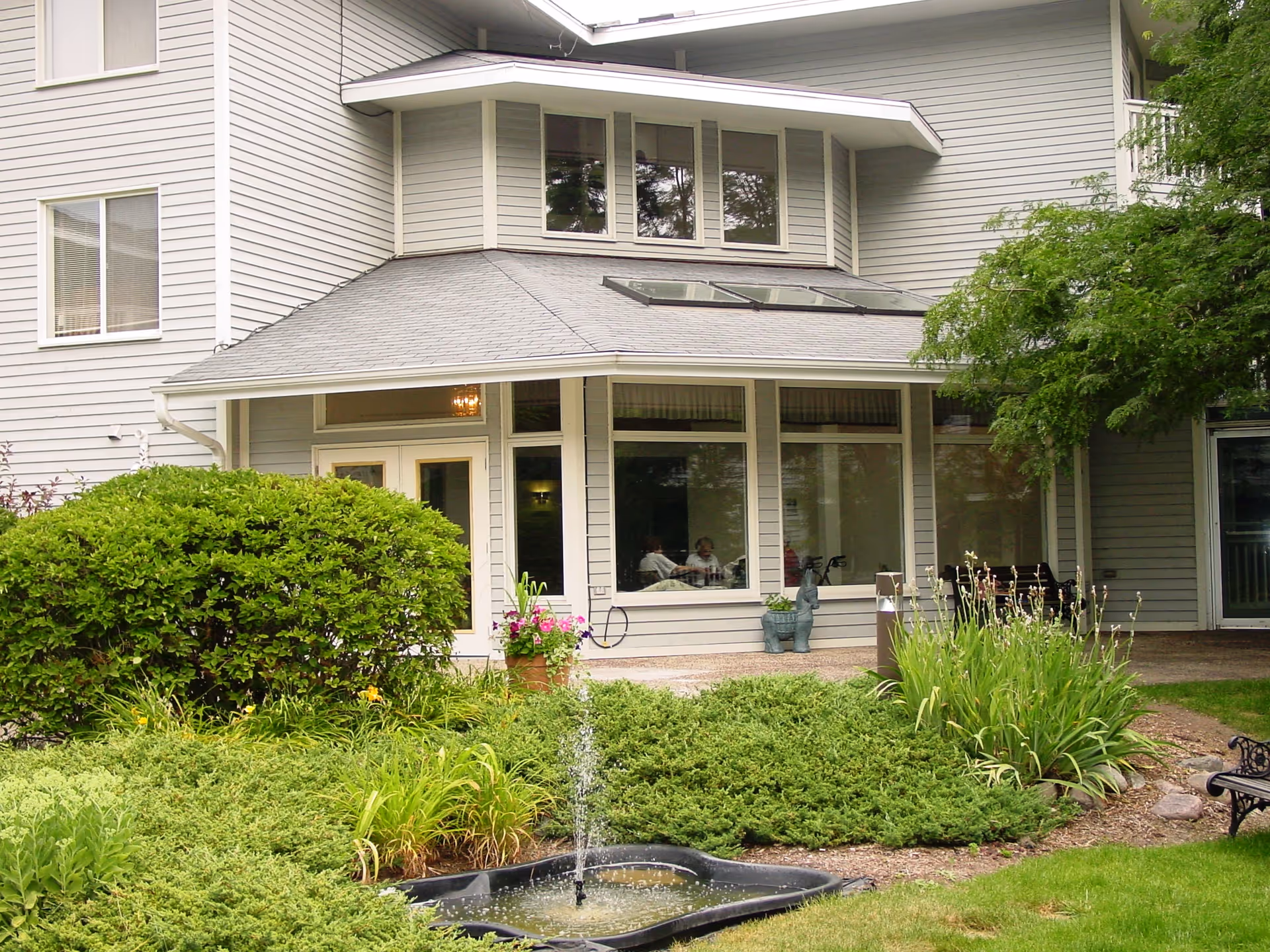 Exterior view of a senior living facility building with gray siding, large windows, and a small fountain surrounded by green bushes and plants in the foreground. There is a bench on the right side and some people visible inside through the windows.