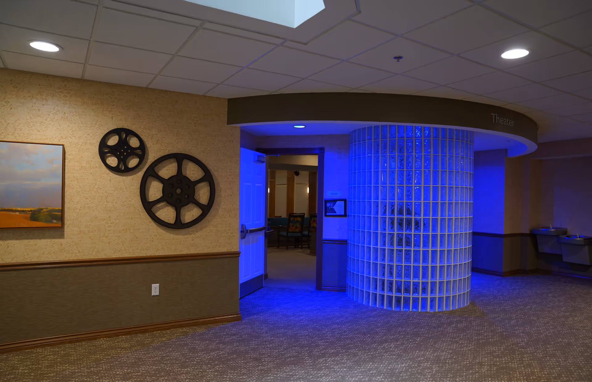 Interior hallway of a retirement community with a curved glass block wall illuminated by blue lighting. The hallway leads to a theater room with an open door showing chairs inside. The wall has two decorative film reels and a framed landscape painting. There are two drinking fountains on the right side of the hallway.
