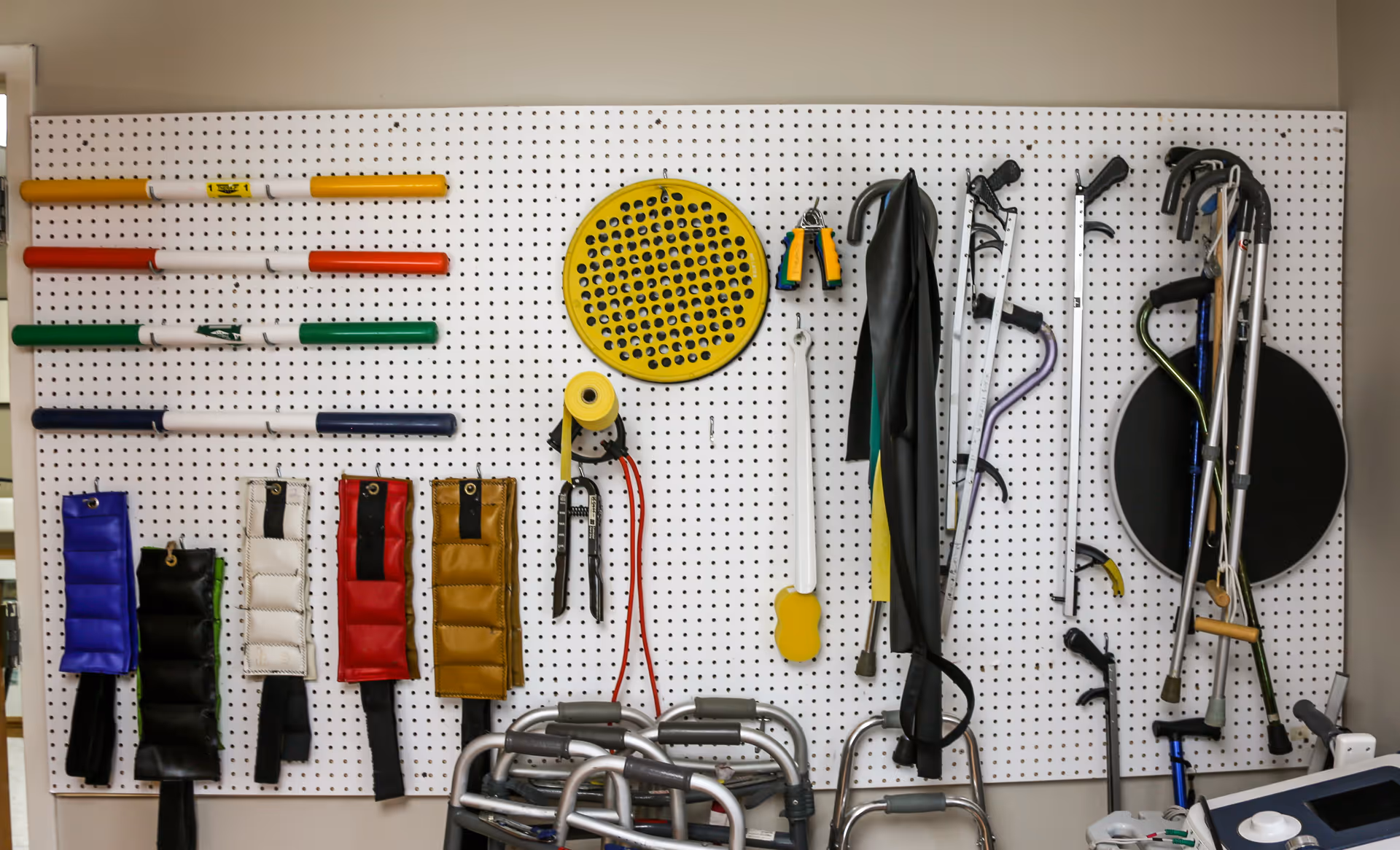 A pegboard mounted on a wall holding various physical therapy and mobility equipment including colored weighted bars, ankle weights, hand grips, a yellow circular pegboard, a hand exerciser, crutches, canes, and walkers.