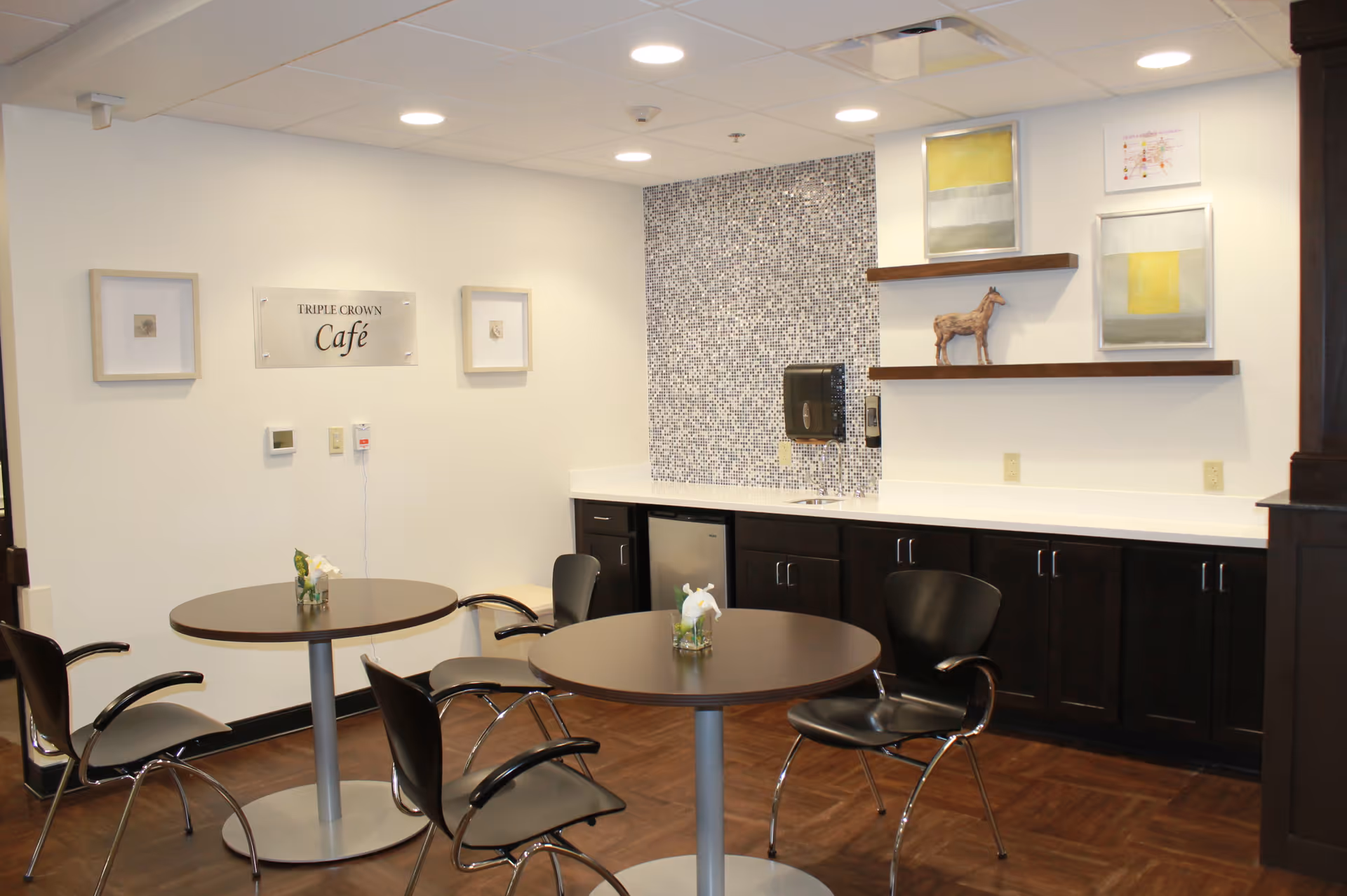 Small cafe seating area with round tables and chairs in front of a counter, mosaic backsplash, and a 'Triple Crown Café' sign.