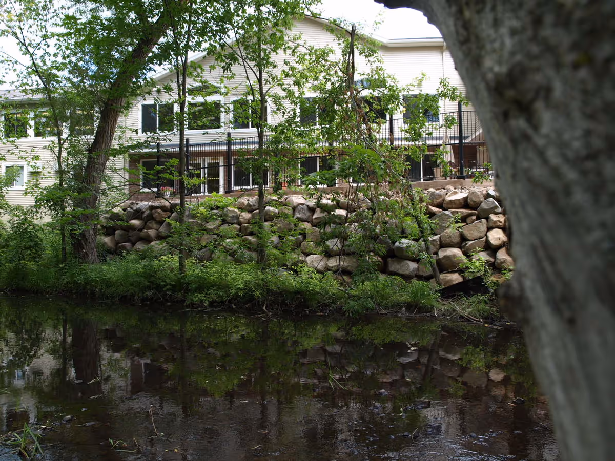 View of a light-colored building partially obscured by trees and a stone retaining wall, with a calm body of water in the foreground reflecting the greenery and structure.