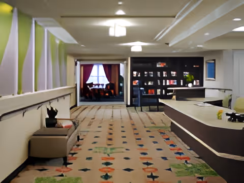 Interior view of a senior living facility hallway with patterned carpet, a bench on the left side, and a reception desk on the right. At the end of the hallway, there is a room with a window covered by curtains and some furniture visible inside.