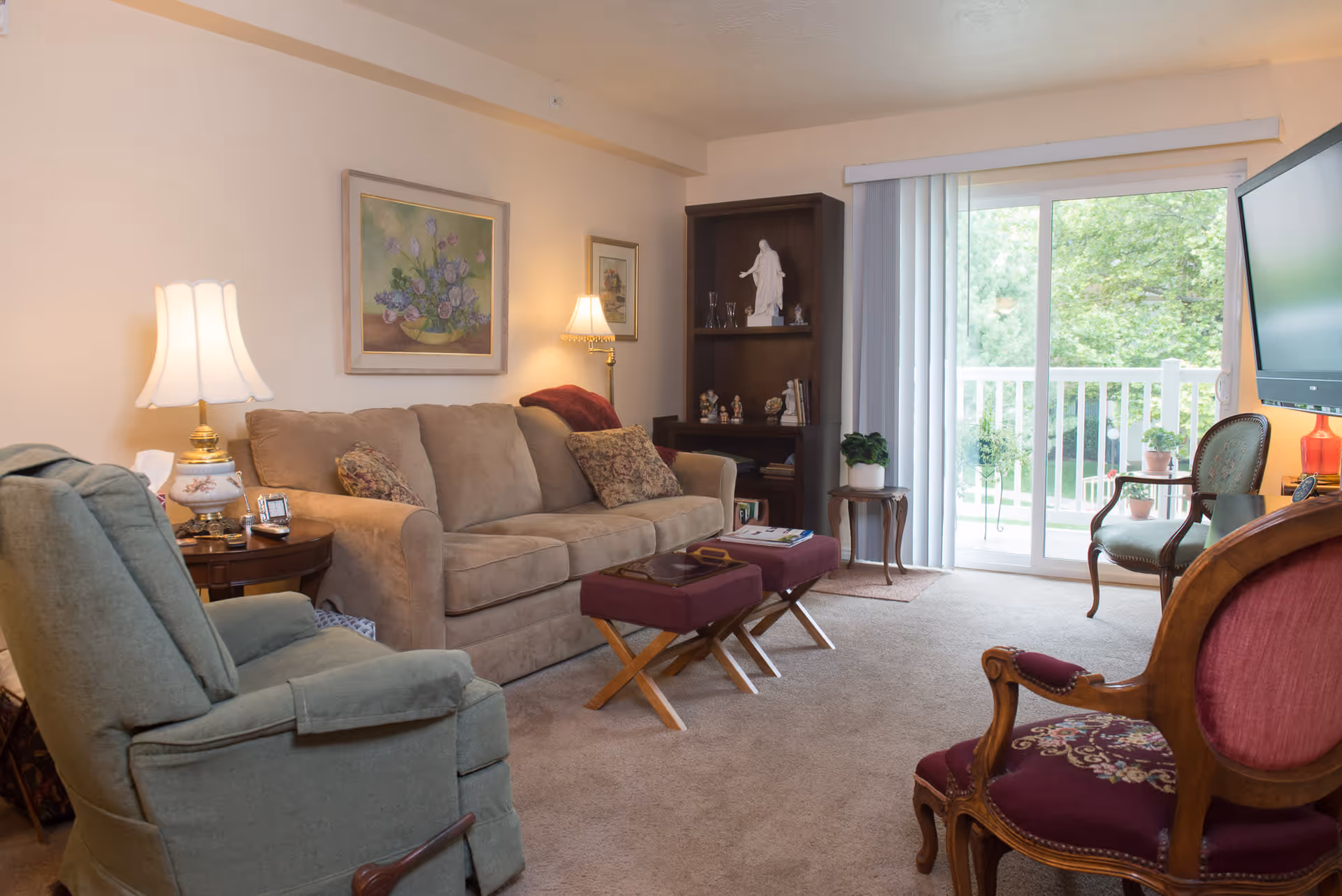 A cozy living room in a retirement facility featuring a beige sofa with patterned cushions, a green recliner chair, two purple cushioned stools, and a wooden armchair with red upholstery. The room has a wooden bookshelf with decorative items, a side table with a lamp, a large flat-screen TV mounted on the wall, and a sliding glass door leading to a balcony with greenery outside.