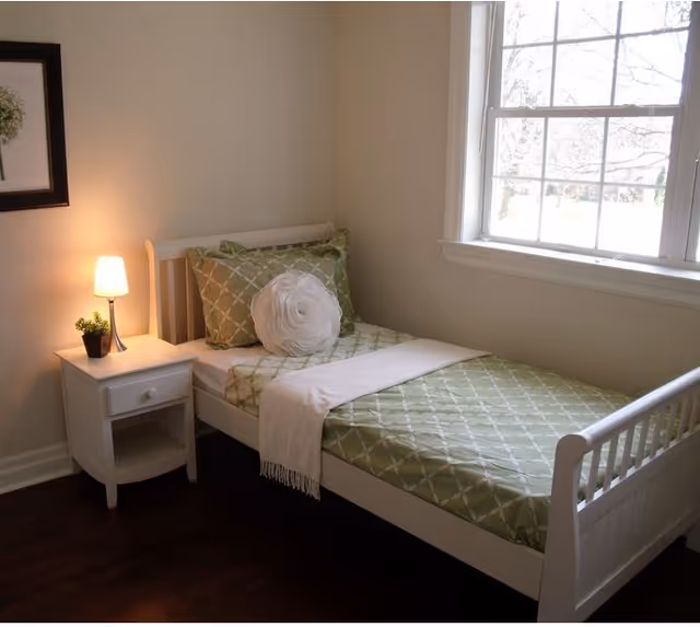 A small bedroom with a single white bed featuring green patterned bedding and a round white decorative pillow. Next to the bed is a white nightstand with a small lamp and a potted plant. A large window lets in natural light, and a framed picture hangs on the wall above the nightstand.