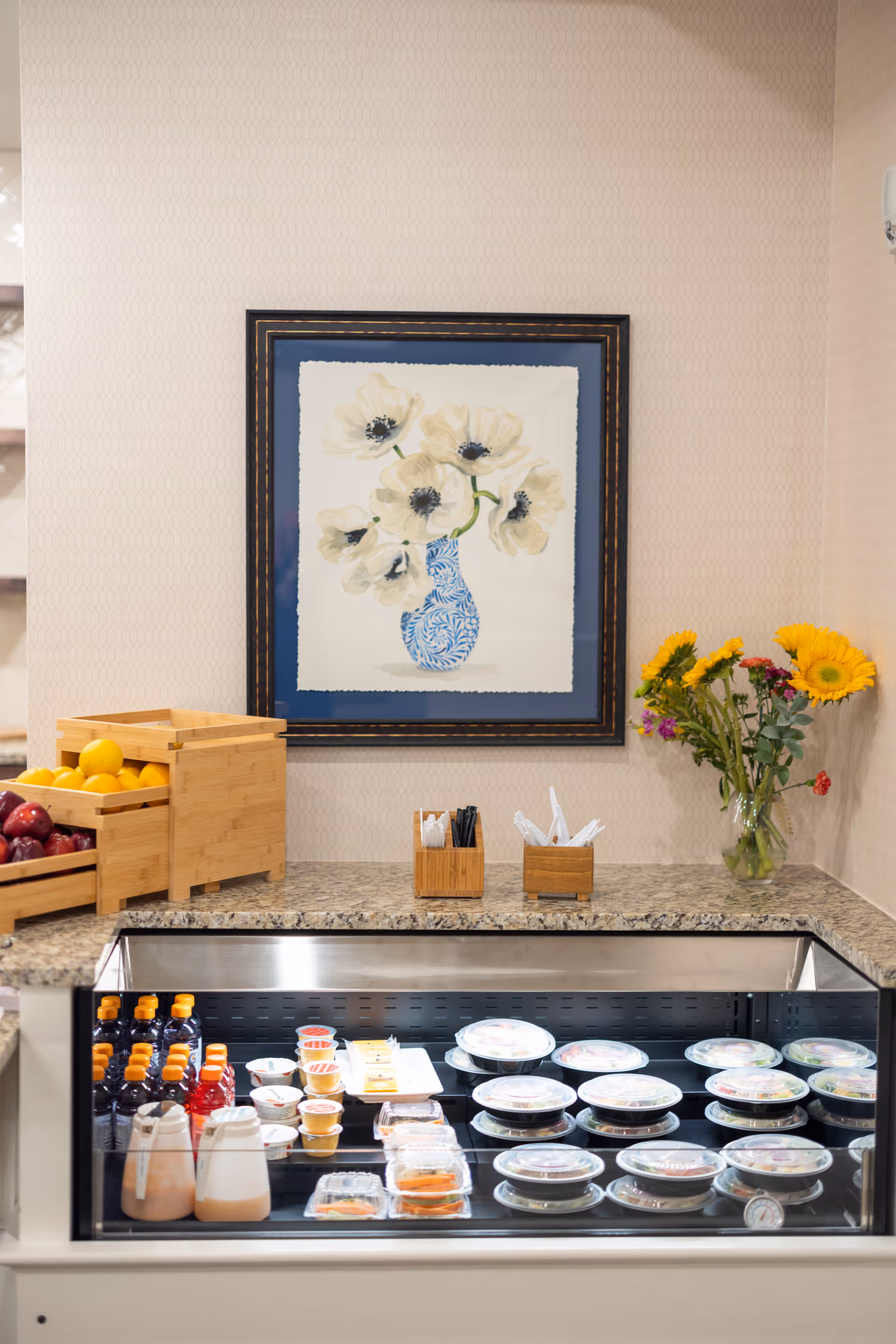 A countertop with a refrigerated display case containing various packaged food items and beverages. On the counter are wooden containers holding utensils and napkins, a vase with colorful flowers, and wooden crates filled with apples and oranges. Above the counter hangs a framed painting of white flowers in a blue vase.
