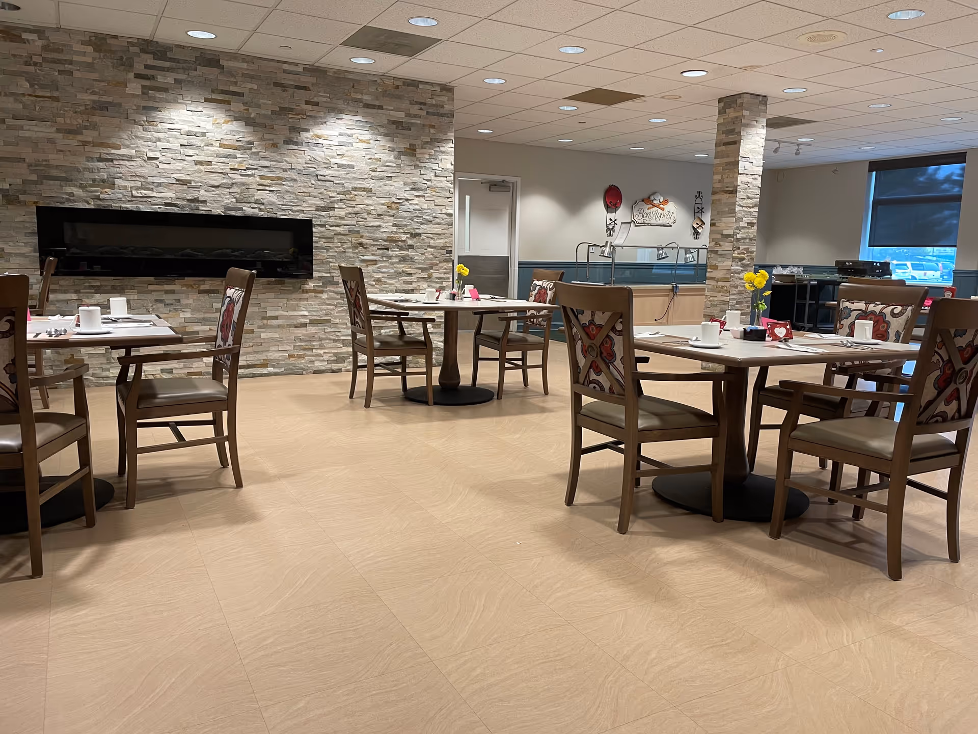 A dining room with several wooden tables and chairs, each table set with cups, napkins, and small flower vases. The room features a stone accent wall with a modern horizontal fireplace and a pillar with matching stone. There is a buffet area in the background with a 'Bon Appetit' sign on the wall and a window with a dark roller shade.