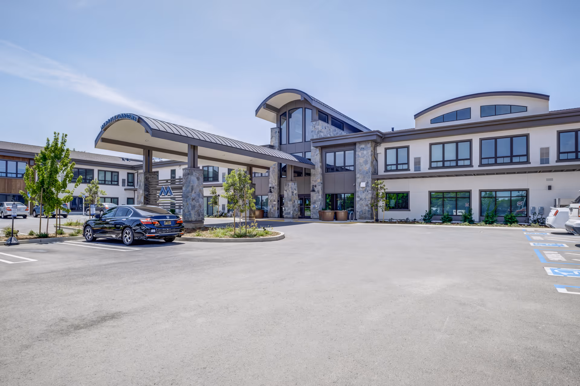 Exterior view of Merrill Gardens at Brentwood senior living facility showing the main entrance with a covered drop-off area supported by stone pillars, a parking lot with several cars, and a clear blue sky.