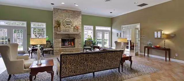 Sunlit communal living room with sofas and armchairs arranged around a central stone fireplace and large windows.