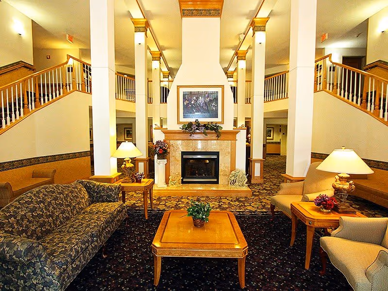 A spacious and well-lit living room area in a retirement community featuring a central fireplace with a framed picture above it. The room has a patterned carpet, a floral upholstered sofa, two armchairs, wooden side tables with lamps, and a wooden coffee table with a small plant. The room also has white pillars and a staircase with wooden railings on both sides.