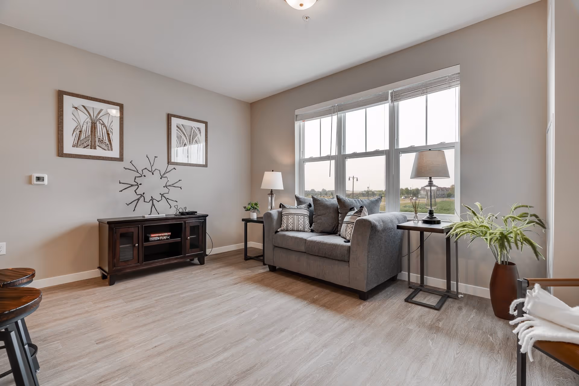 A bright and modern living room with a gray loveseat adorned with patterned pillows, two side tables each holding a lamp, a wooden media console with decorative items, two framed architectural photos on the wall, large windows letting in natural light, and a potted plant in the corner.