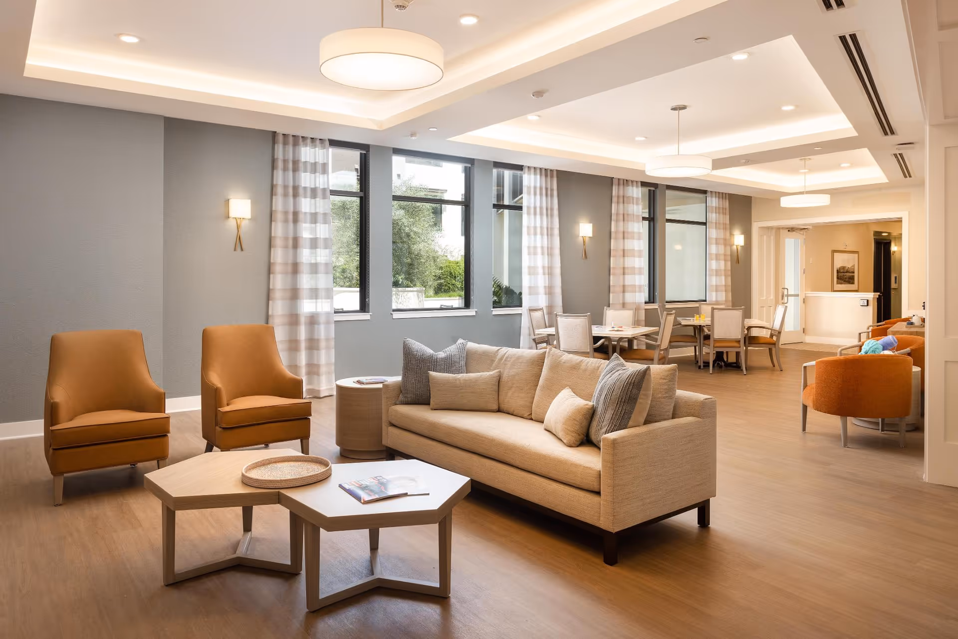 A spacious and well-lit senior living common area with a beige sofa adorned with multiple cushions, two orange armchairs, and two hexagonal wooden coffee tables. The room features large windows with striped curtains, light gray walls, wooden flooring, and modern ceiling lights. In the background, there are dining tables with chairs and additional seating areas.