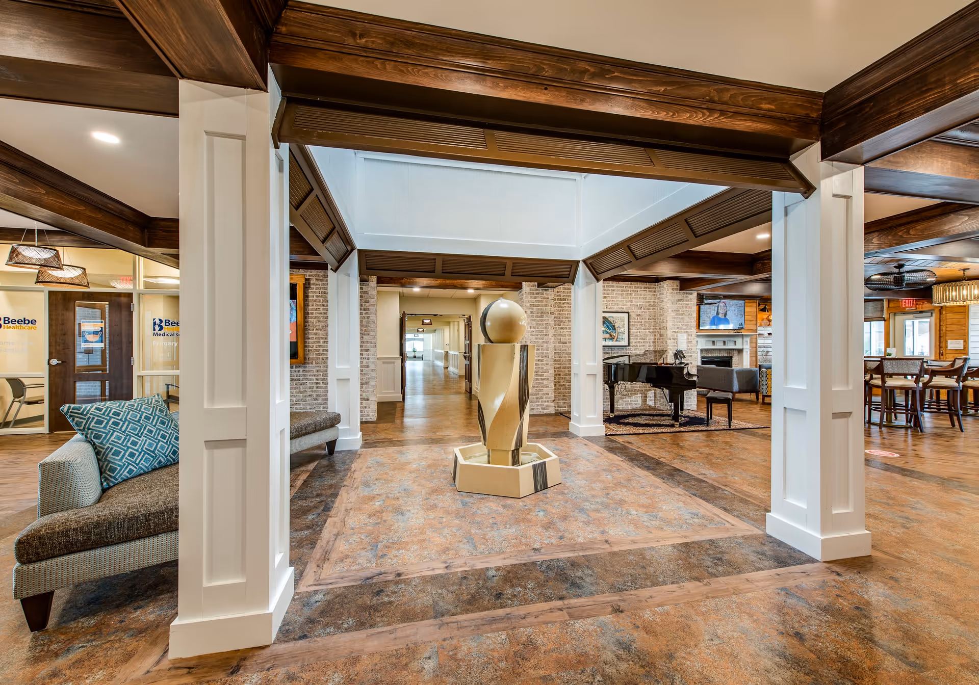 Interior view of a senior living facility lobby area with a modern sculpture in the center, surrounded by white pillars and wood beams. To the left, there is a seating area with a couch and a glass door with Beebe Healthcare signage. To the right, there is a lounge area with a grand piano, a TV mounted above a fireplace, and a dining area with tables and chairs. The floor has a wood and tile pattern, and the walls are a mix of brick and painted surfaces.