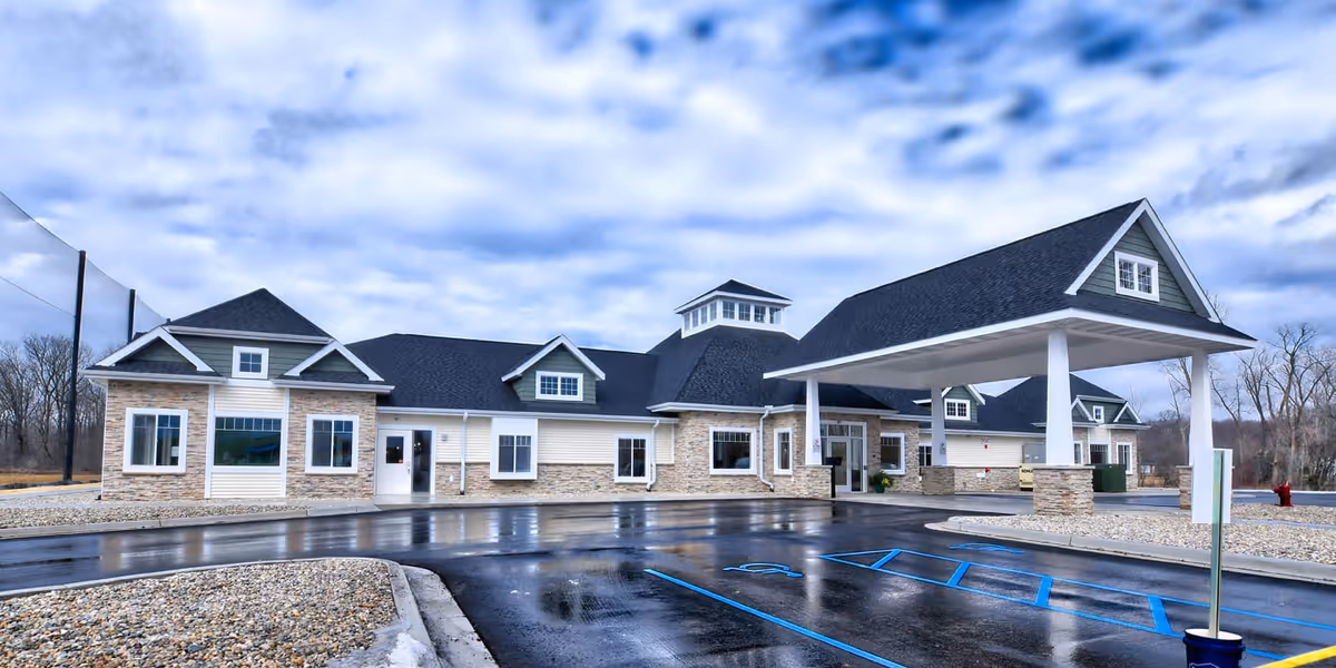 Exterior view of a single-story senior living facility building with stone and siding facade, multiple windows, a covered entrance with white pillars, and a wet parking lot with handicap parking spaces marked in blue. The sky is cloudy and there are leafless trees in the background.