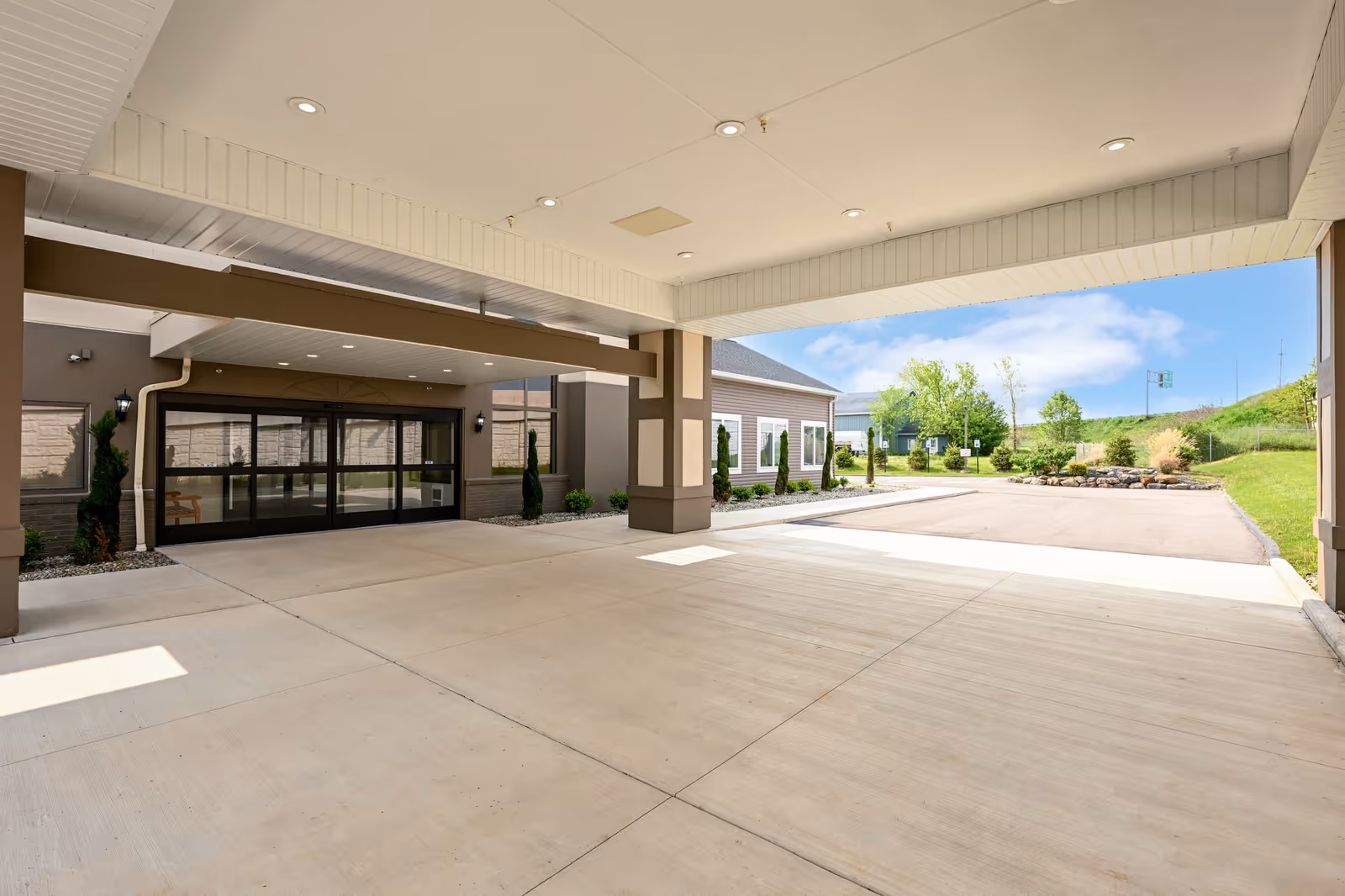 Covered entrance area of Hines Hill Senior Living facility with automatic sliding glass doors, beige and brown exterior walls, small landscaped areas with shrubs, and a clear blue sky in the background.