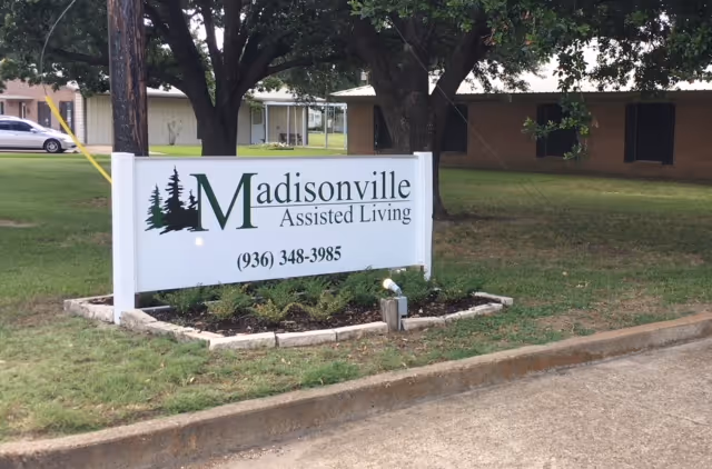 Outdoor view of a sign for Madisonville Assisted Living placed on a grassy area with trees and buildings in the background. The sign includes the facility name and phone number (936) 348-3985.