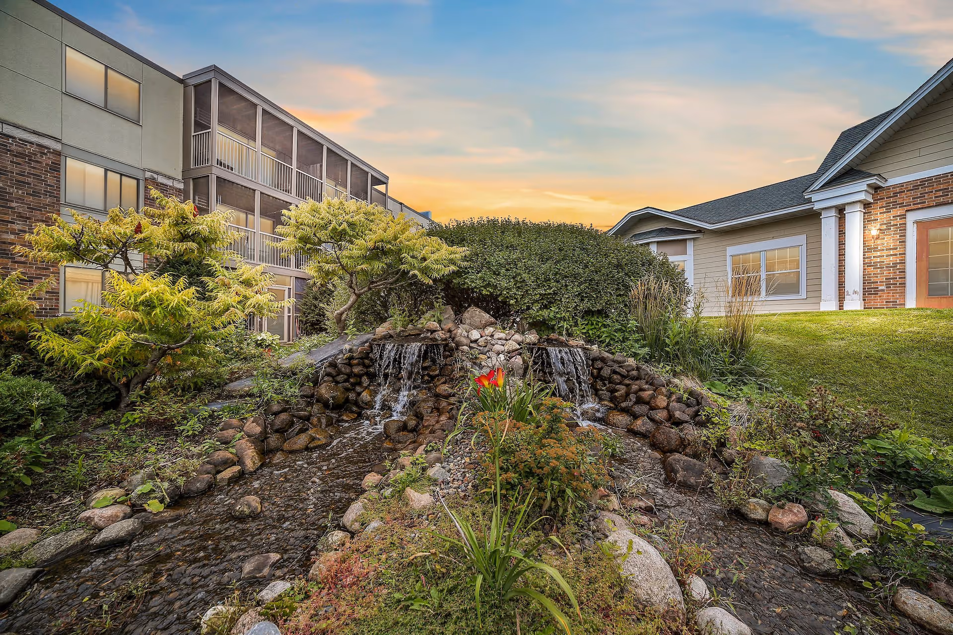 A landscaped outdoor area at the Waterford facility featuring a small double waterfall flowing over rocks into a shallow stream surrounded by greenery and flowering plants, with buildings visible in the background under a partly cloudy sky at sunset.
