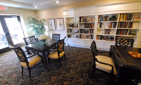 A cozy senior living common area with a dark wooden table surrounded by four chairs with yellow cushions. There is a large built-in white bookshelf filled with books and decorative items against the wall. A large potted plant is near the glass door entrance, and the room has patterned carpet flooring and soft lighting.