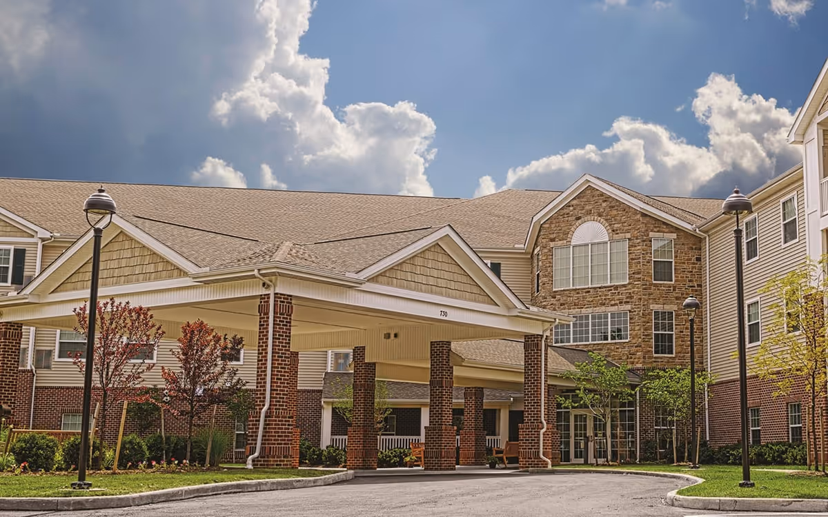 Exterior view of a senior living facility named Danbury Westerville featuring a covered entrance supported by brick columns, surrounded by well-maintained landscaping with small trees and bushes under a partly cloudy sky.