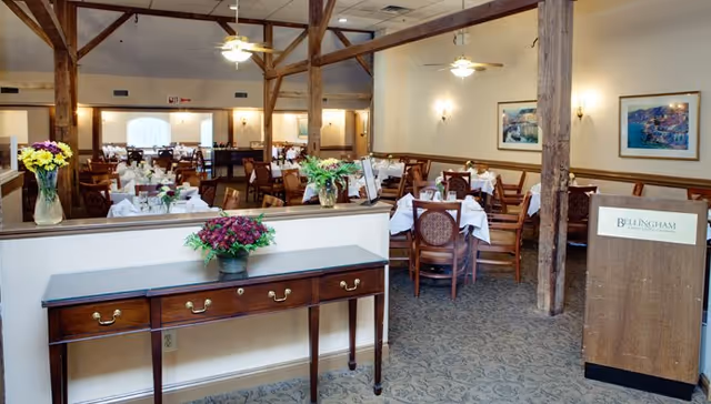 Dining room with multiple set tables, wooden support beams, floral centerpieces, and a podium displaying a Bellingham sign.