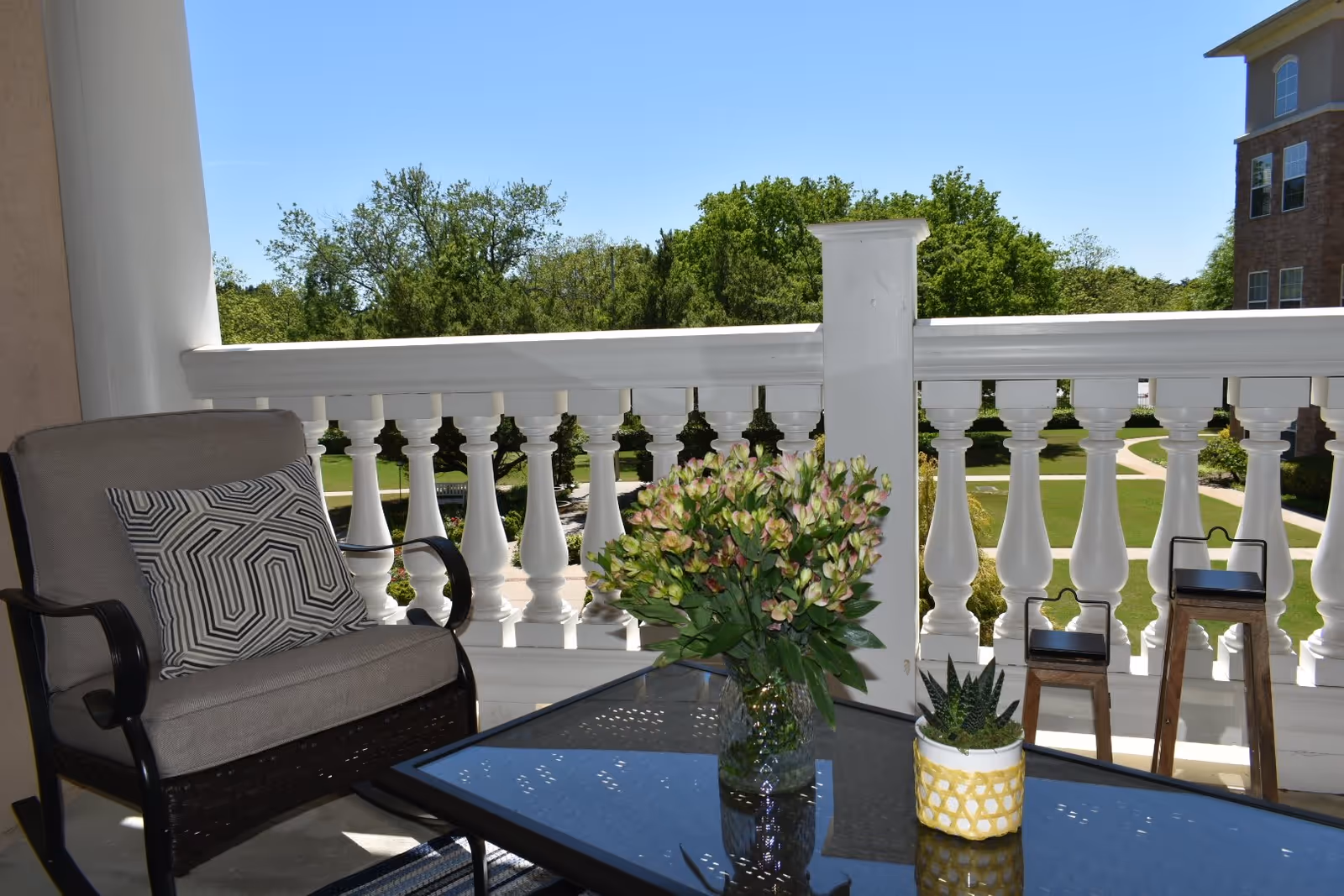 A balcony with a cushioned chair featuring a patterned pillow, a glass-top table with a vase of flowers and a small potted plant, white balustrade railing, and a view of green trees and a building in the background under a clear blue sky.