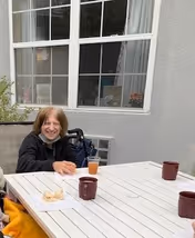 An elderly person sitting outside at a white table with three maroon mugs and a glass of orange drink. The person is smiling and there is a wheelchair behind them. The background shows a gray building wall with a window.